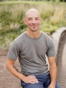 A bald man wearing a gray t-shirt and jeans, sitting outdoors with greenery in the background.