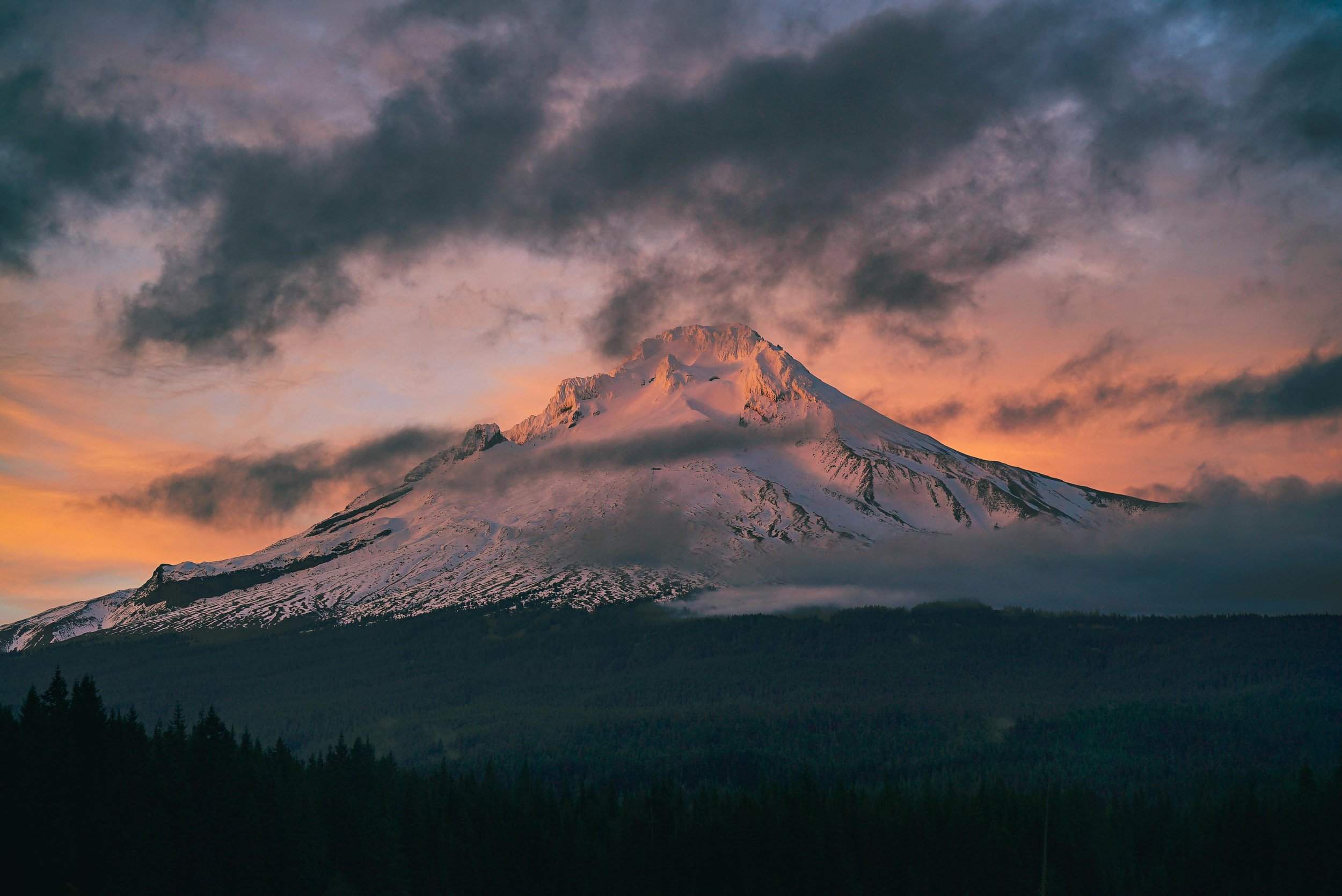Snow-capped mountain, possibly Mount Hood, under a colorful sunset sky with dark clouds and a forested landscape in the foreground.