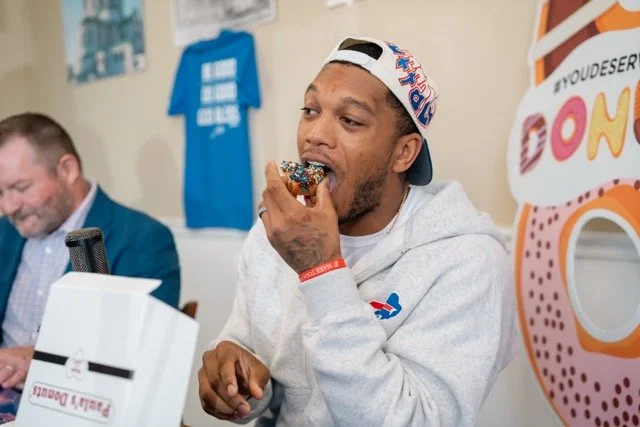 Man eating a donut with colorful sprinkles at an event, wearing a cap backwards and a gray hoodie, with a man in a suit in the background and themed decorations.