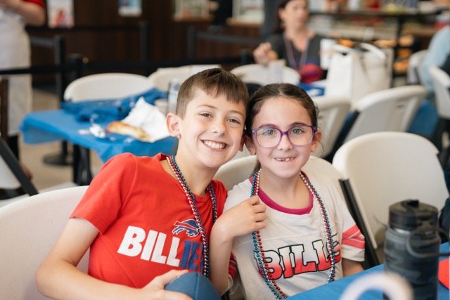 Two children, a boy and a girl, smiling and sitting close together at a party, both wearing festival beads and themed shirts, with a busy event setting in the background.