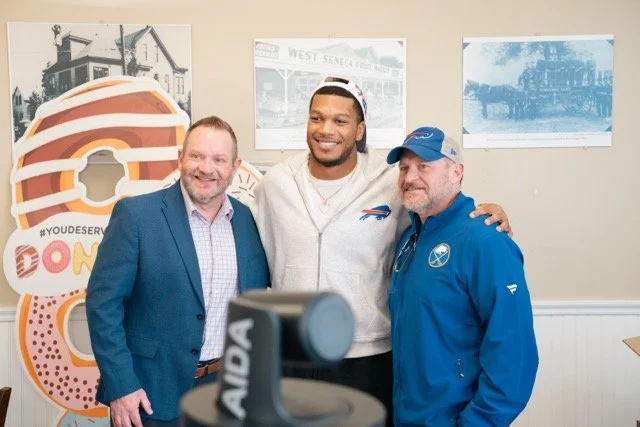 Three men standing together in a room, smiling for a photo. One in a blue suit, one in a white hoodie with a cap, and another in a blue jacket with a cap. There is a large donut-shaped sign and framed pictures on the wall behind them.