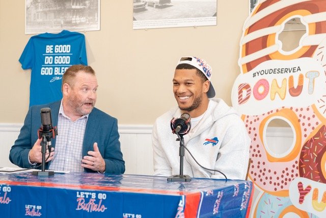 Two men seated at a table engaged in a conversation, with microphones in front of them. The man on the left is wearing a blue blazer and a checked shirt, the man on the right is wearing a white hoodie and a backwards baseball cap. Behind them is a la