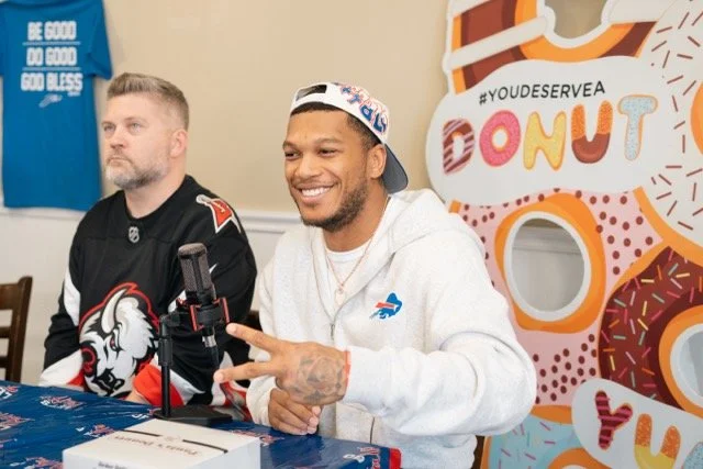 Two men sitting at a table with a microphone, one smiling and holding up a peace sign, in front of a colorful donut-themed backdrop.