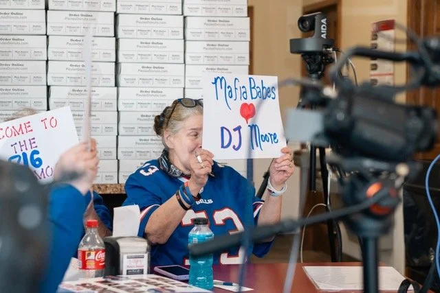 A woman wearing a blue sports jersey with the number 3 is laughing and holding a sign that says "Maifa Babes DJ Moore" and has a red heart on it. She appears to be at a table with water bottles and other items, and there is a camera pointed at her.