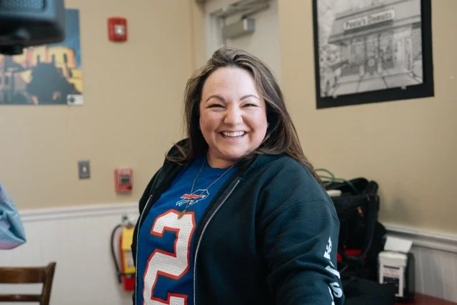 A smiling woman with long brown hair wearing a blue sports jersey and a dark jacket in a room with framed pictures on the wall.