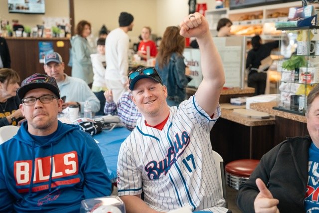 Group of people at a restaurant, including a man in a Buffalo Bills jersey raising his fist and smiling, and another man in a blue hoodie with a 