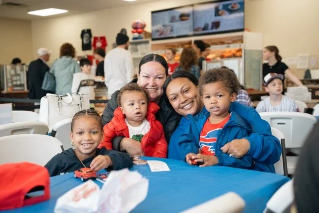 A group of smiling women and children sitting at a table in a busy cafeteria or food court.