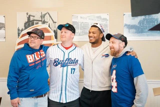 Four men standing together in front of a wall with black and white photos, wearing Buffalo Bills apparel and caps, smiling and posing for the photo.