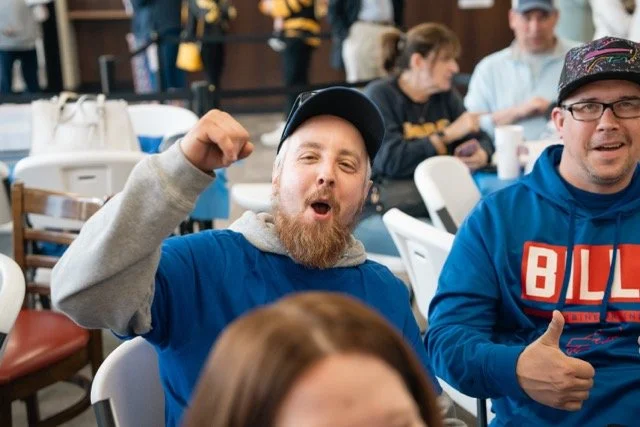A man with a beard and cap making a fist pump gesture at an indoor gathering, surrounded by other people seated at tables.