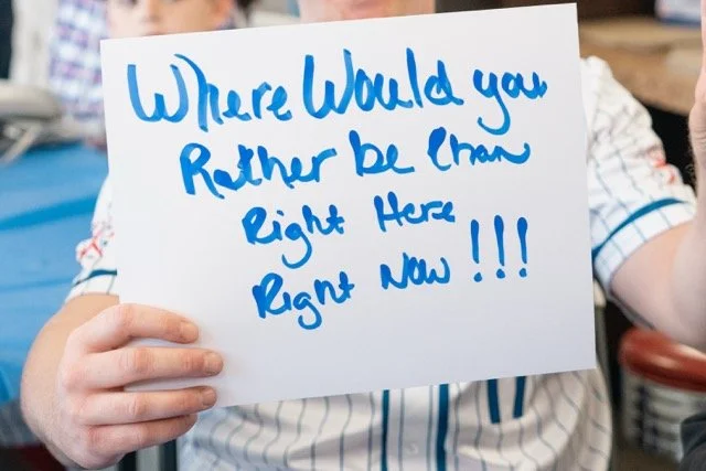 Person holding a white sign with blue handwritten text that says, 'Where would you rather be right now, right here, right now!!!'