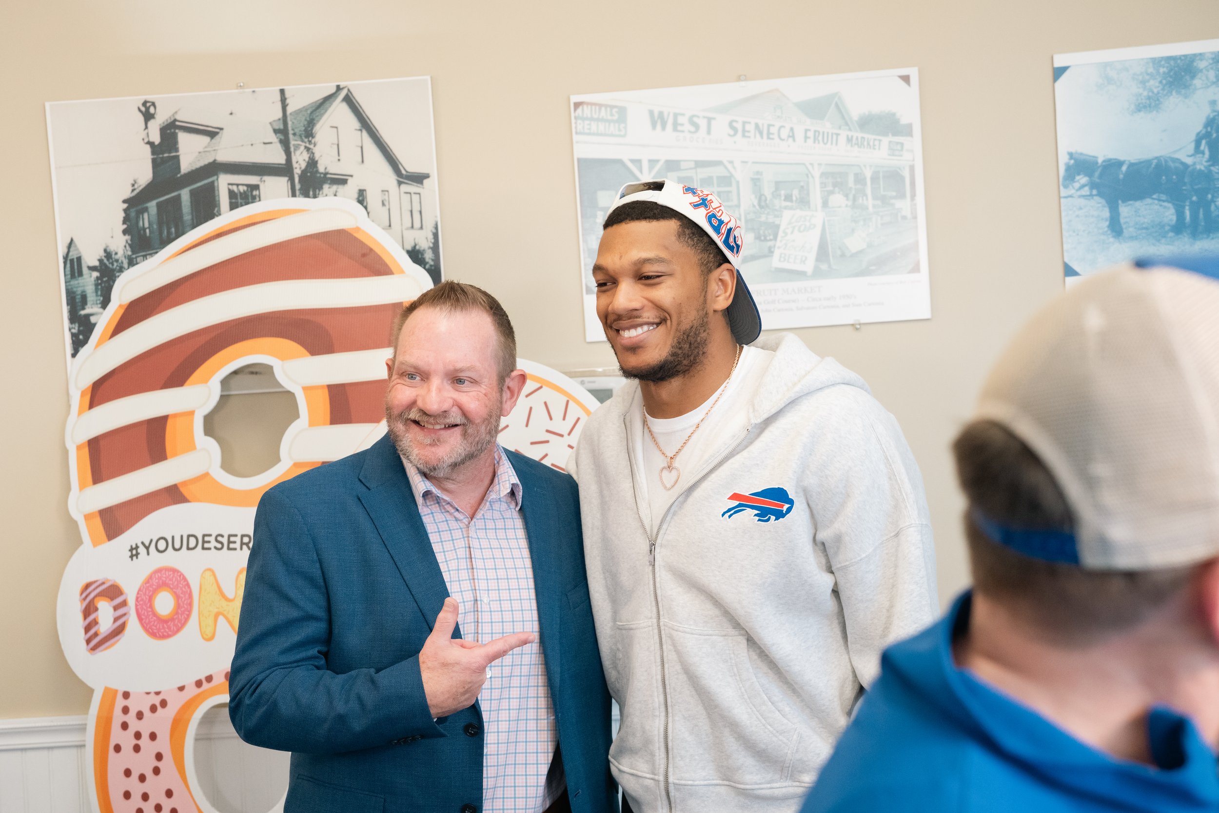 Two men smiling for a photo, one wearing a suit and the other in a hoodie with a sports team logo, with a donut-themed cardboard cutout and vintage-style posters on the wall behind them.