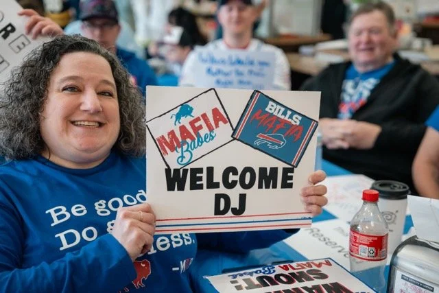 A woman holding a sign that says "Mafia Babies Welcome DJ" at an indoor gathering with other smiling people in the background.