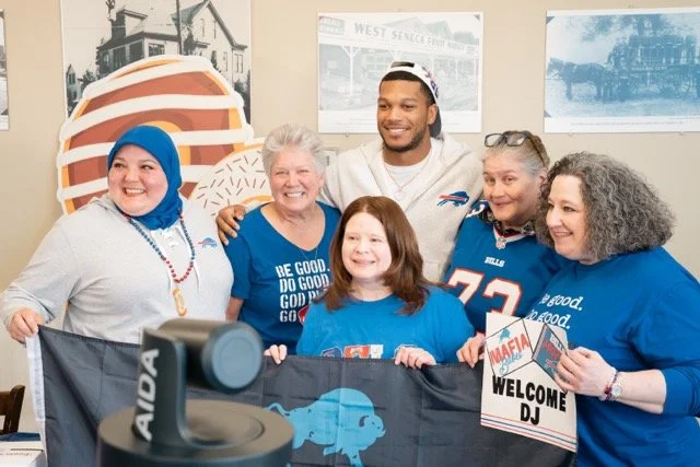 A group of six diverse people, including two children and four adults, smiling and posing together at a fan event, with a football-themed backdrop and a sign that says 'Welcome DJ'.