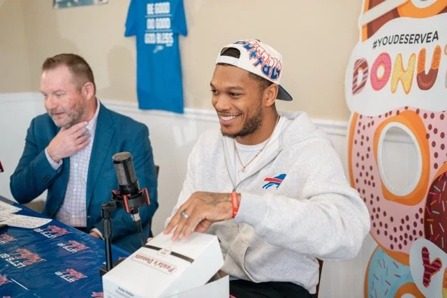 A man in a white hoodie and colorful snapback hat smiling as he opens a box at a table, with a man in a blue blazer beside him. There is a donut-themed banner in the background.