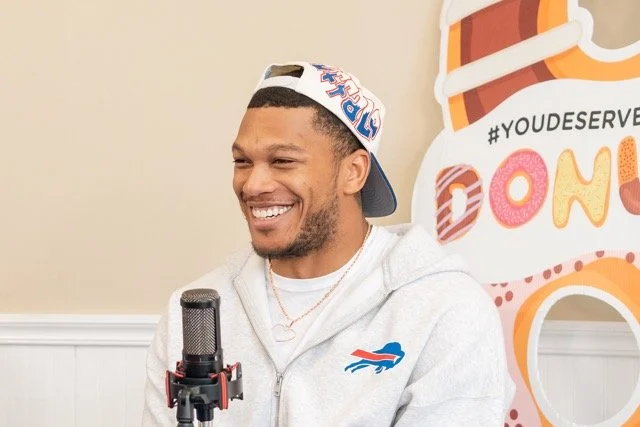 A man smiling, wearing a backward baseball cap and a gray hoodie with a Buffalo Bills logo, sitting in front of a microphone with a colorful backdrop that says "#YOU DESERVE" and "DONUT."