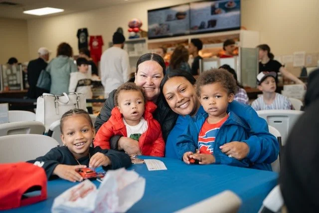 A group of children and two women smiling and sitting at a table in a busy indoor setting, likely a restaurant or cafe, with a menu and people in the background.
