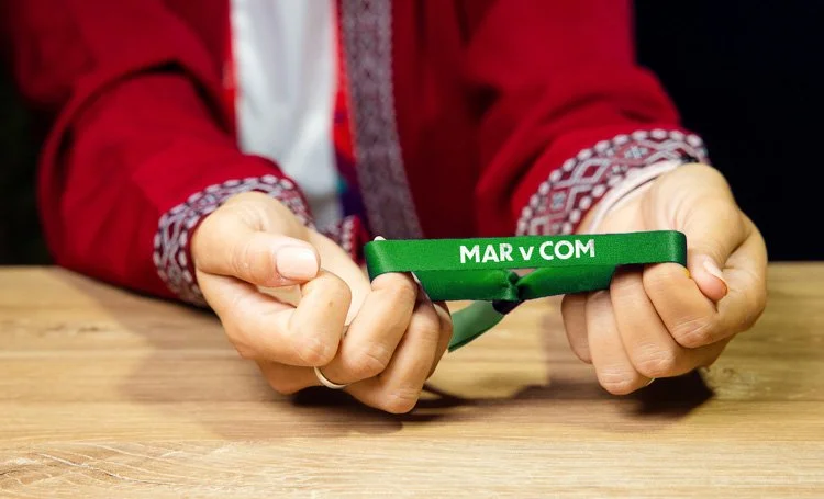 Person in a red sweater holding a green wristband with the words "MAR v COM" on it, resting on a wooden table.
