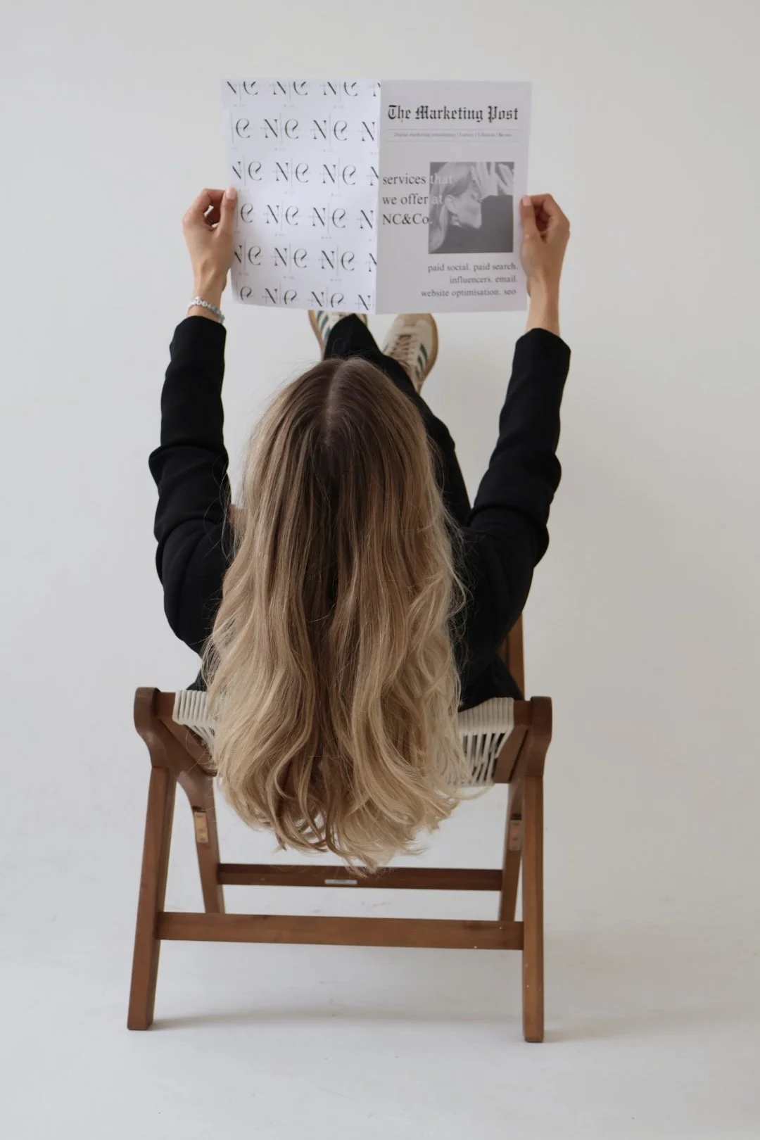 A woman with long, wavy blonde hair sitting on a wooden chair, viewed from above, holding a printed paper with various texts and images.