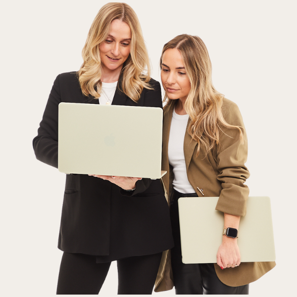 Two women looking at a laptop together against a white background, one holding a laptop and the other holding a tablet.