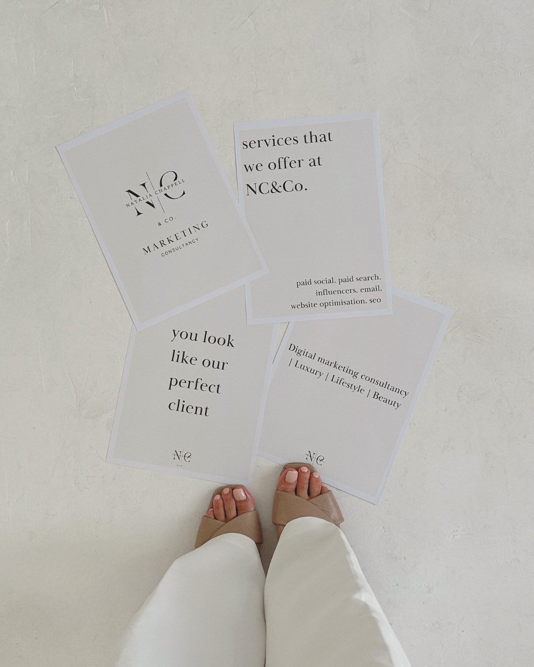 Photo showing a person's feet in beige shoes and white pants, standing on a white floor surrounded by four printed informational flyers.