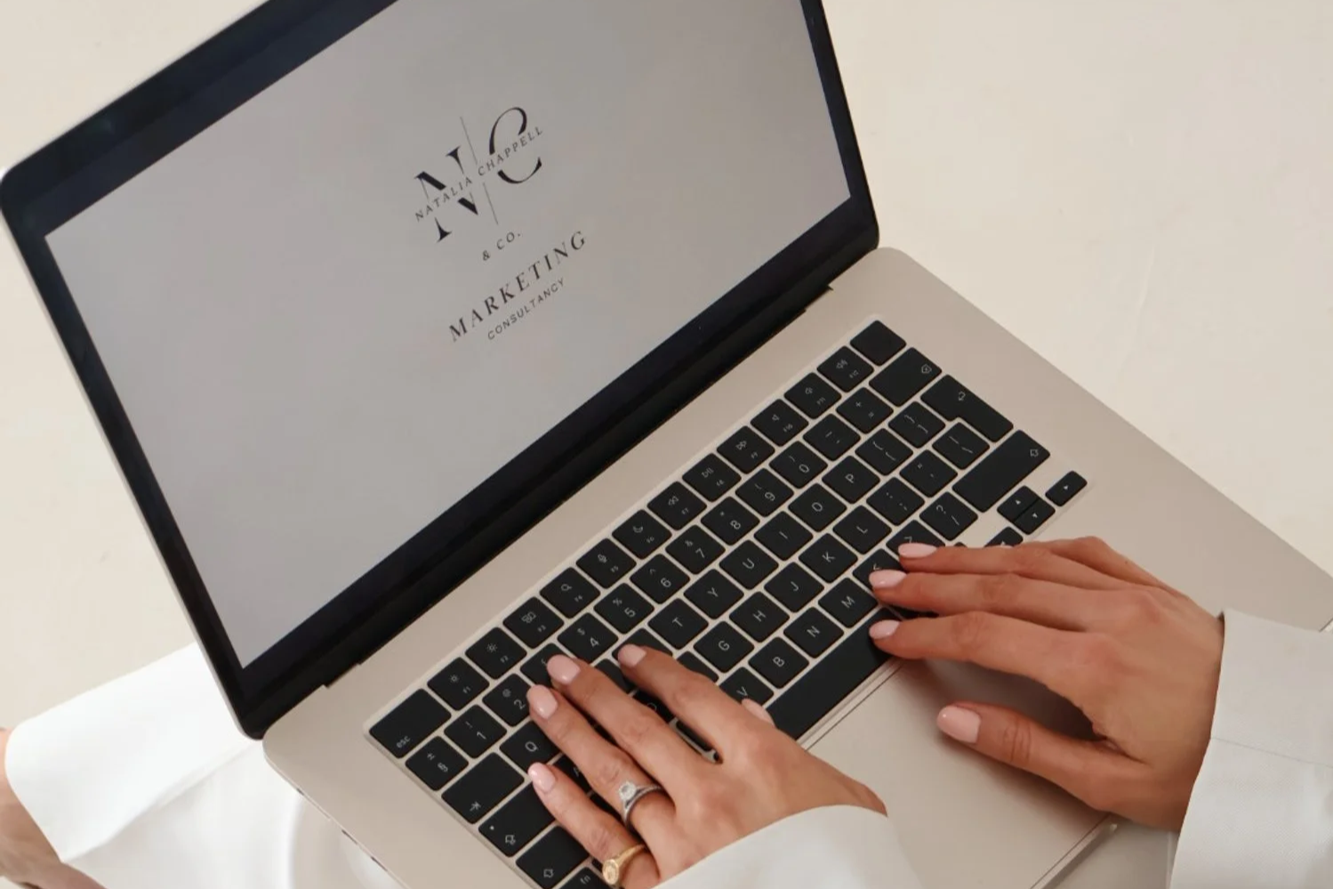 Person typing on a silver laptop with a marketing consultant logo displayed on the screen.