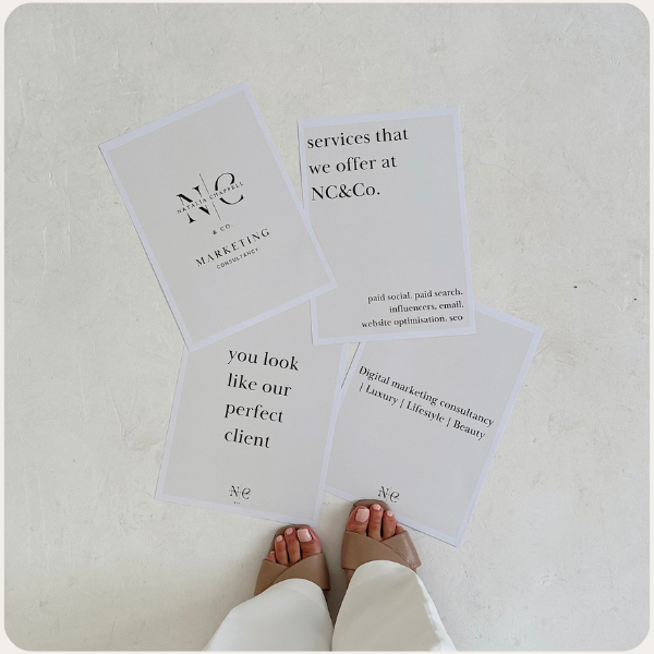 Four printed marketing flyers on a light-colored surface, with a person's feet visible, wearing beige sandals and white pants.