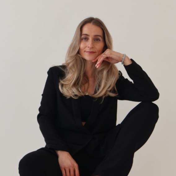 A woman with long blonde hair sitting against a plain white background, wearing a black blazer and pants, resting her chin on her right hand with a slight smile.