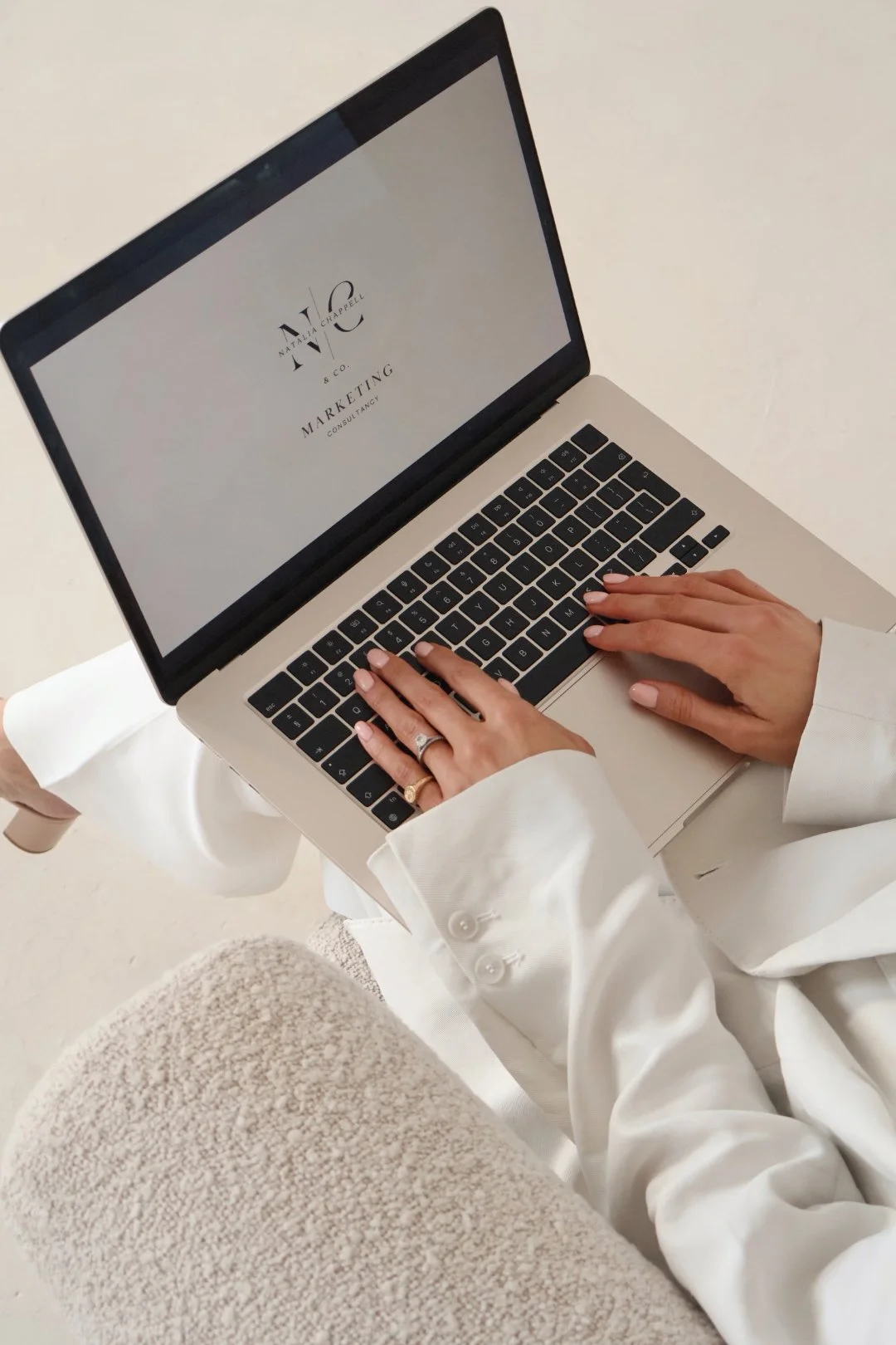 A person wearing a white blazer and beige pants working on a laptop with a screen displaying 'NAT 'N' CHAPEL & CO. MARKETING CONSULTANCY'.