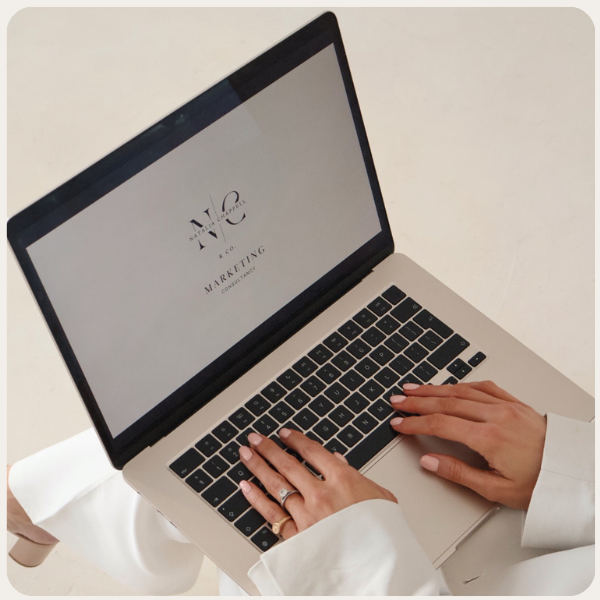 Person typing on a silver laptop with a white and black keyboard, displaying a logo for a marketing company on the screen.