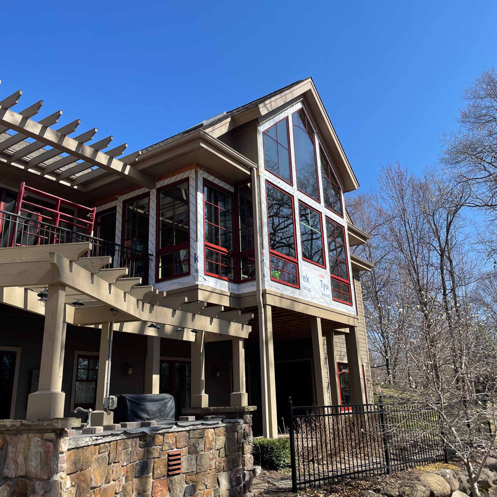 A multi-story house under construction with large windows and beige siding, surrounded by leafless trees, a stone wall, and a black metal fence, with a clear blue sky overhead.