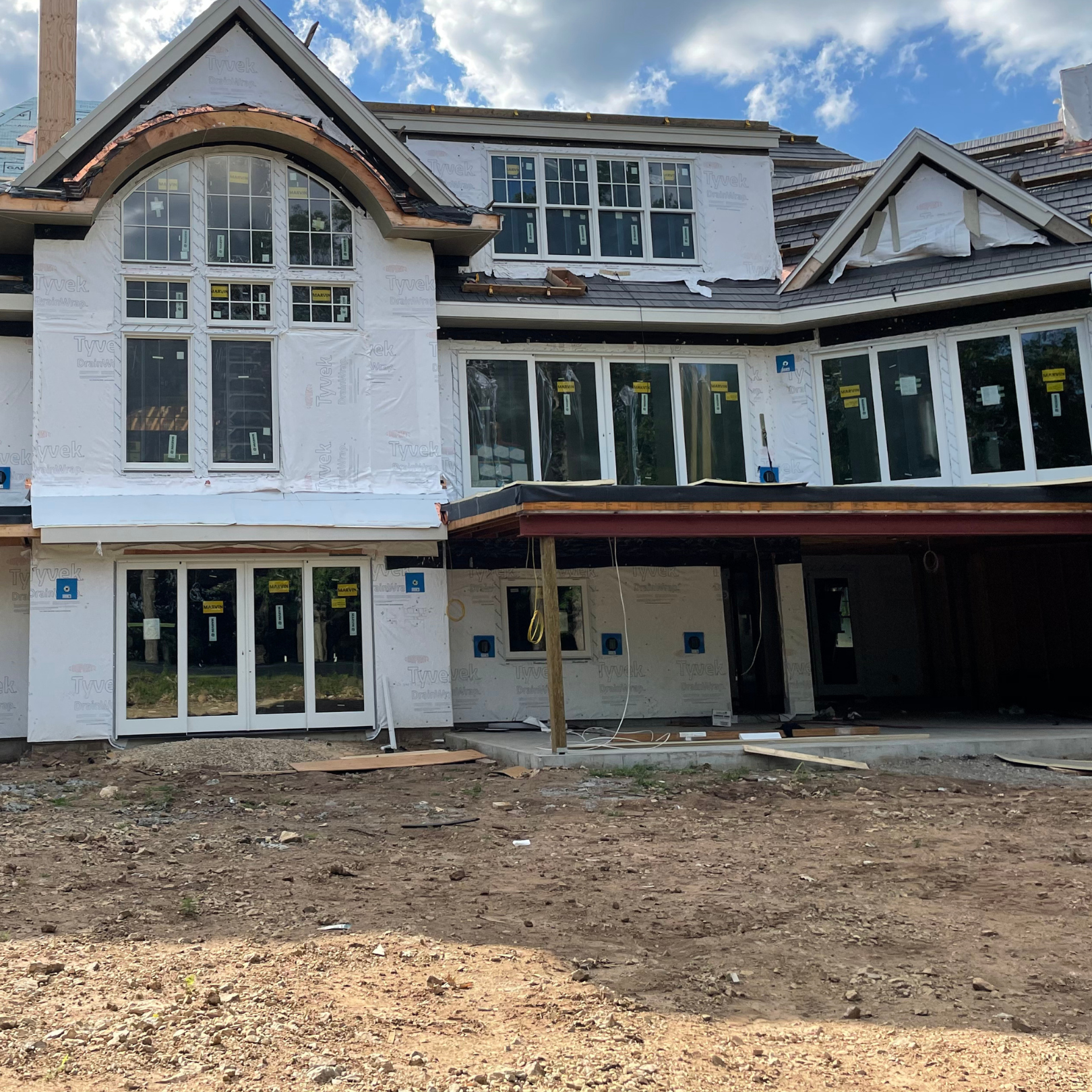 Under construction multi-story house with white window frames and partially installed roofing, surrounded by dirt and construction materials.
