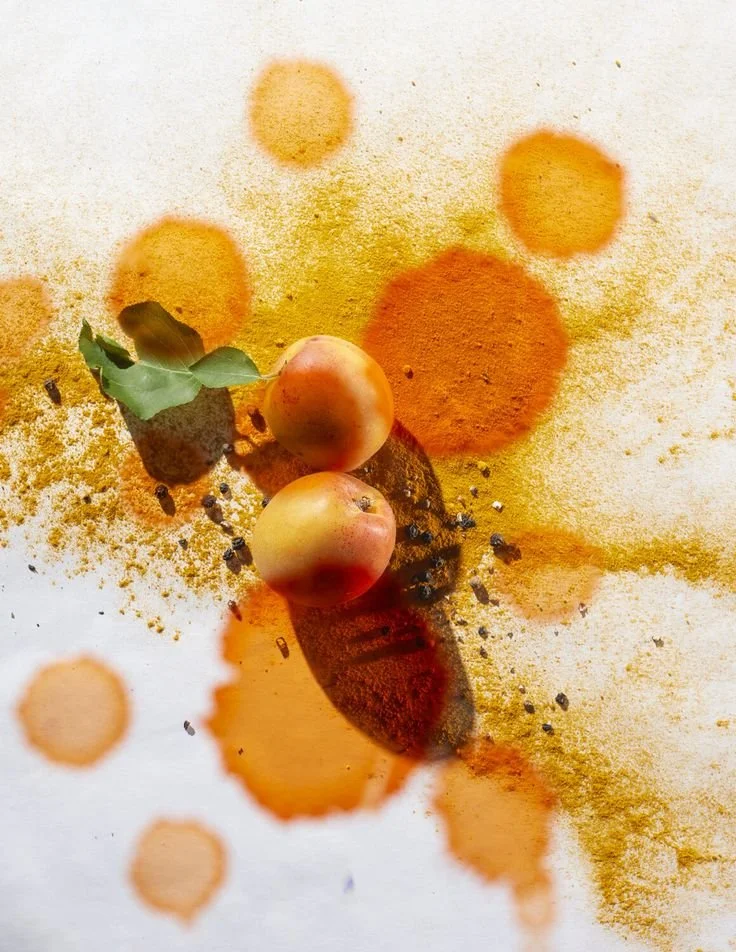 Two peaches with leaves on top of a white surface, surrounded by orange and yellow powder and splashes of orange liquid.