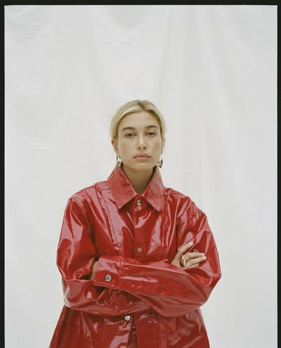 A woman with blonde hair and hoop earrings wearing a shiny red leather jacket, standing with arms crossed against a plain white background.