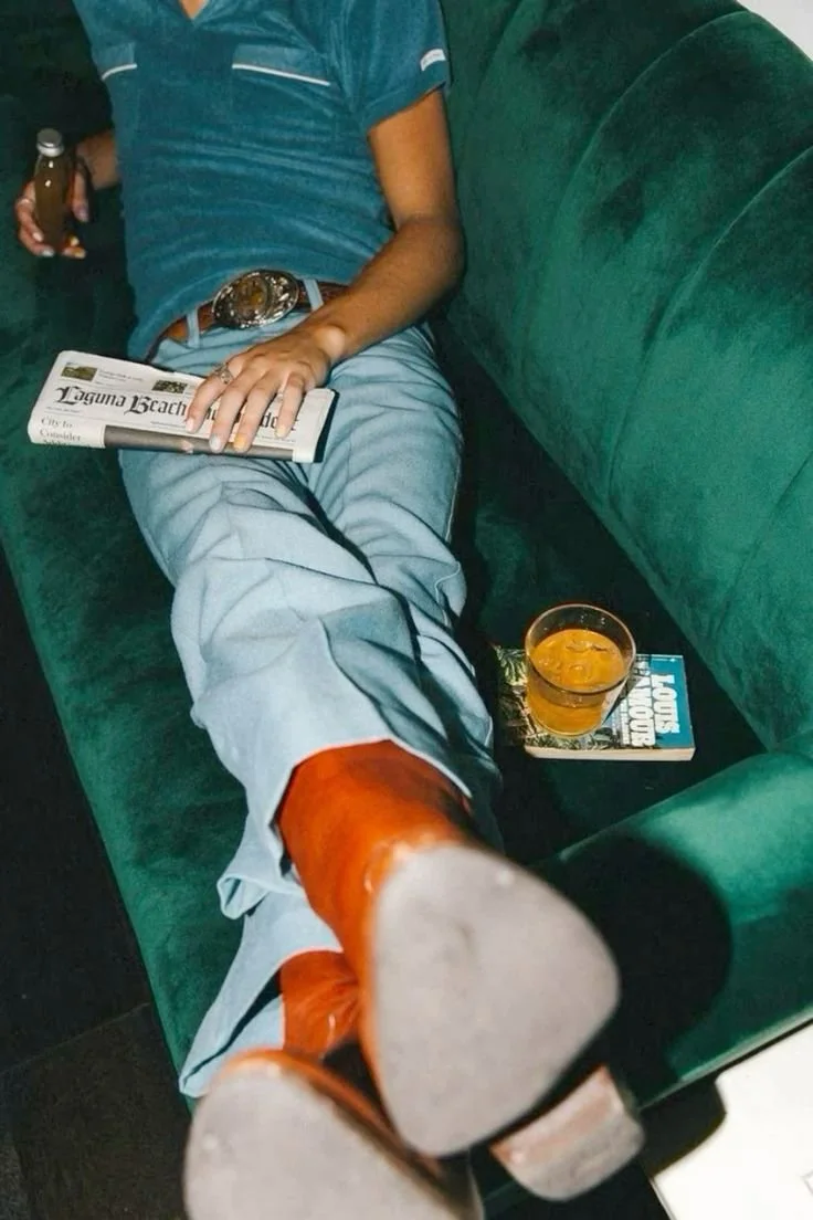 A person reclining on a green velvet couch, wearing a blue shirt, light-colored pants, and orange boots, with one leg stretched out and foot toward the camera. They hold a newspaper in one hand and a drink in the other, with a glass of orange-colored