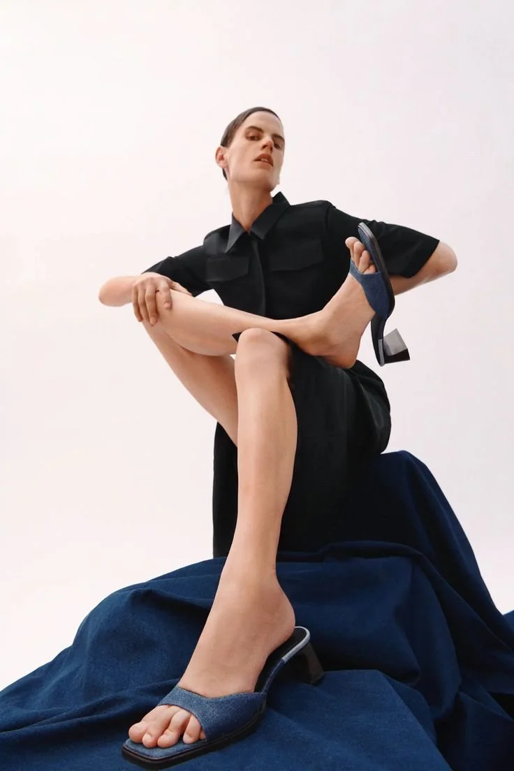 A model in a black uniform, possibly a police officer, sits on a dark fabric covered platform against a white background. The model has one leg raised, showing their foot and sandal, while the other arm is bent, holding their raised leg. The model ha