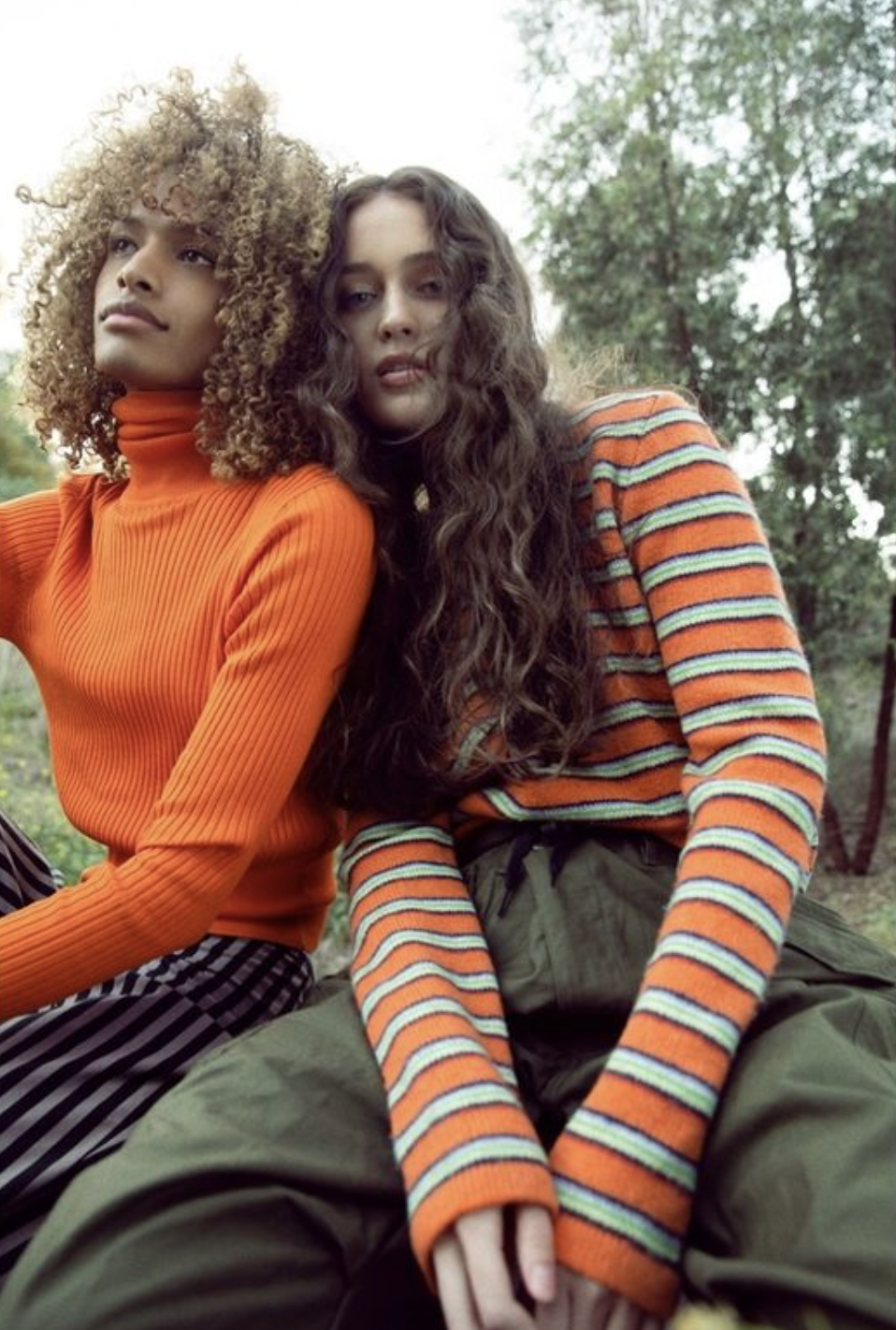 Two women sit outdoors among trees, one with curly blonde hair wearing an orange turtleneck, and the other with long wavy brown hair wearing a colorful striped sweater.