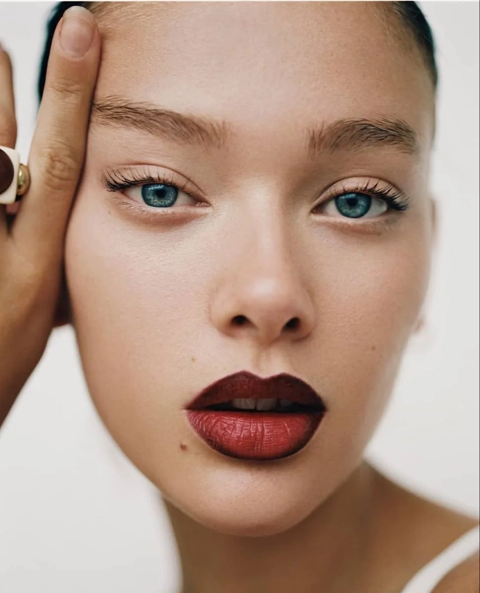 Close-up of a woman with striking blue eyes, dark red lipstick, and light skin, softly touching her forehead with her hand.
