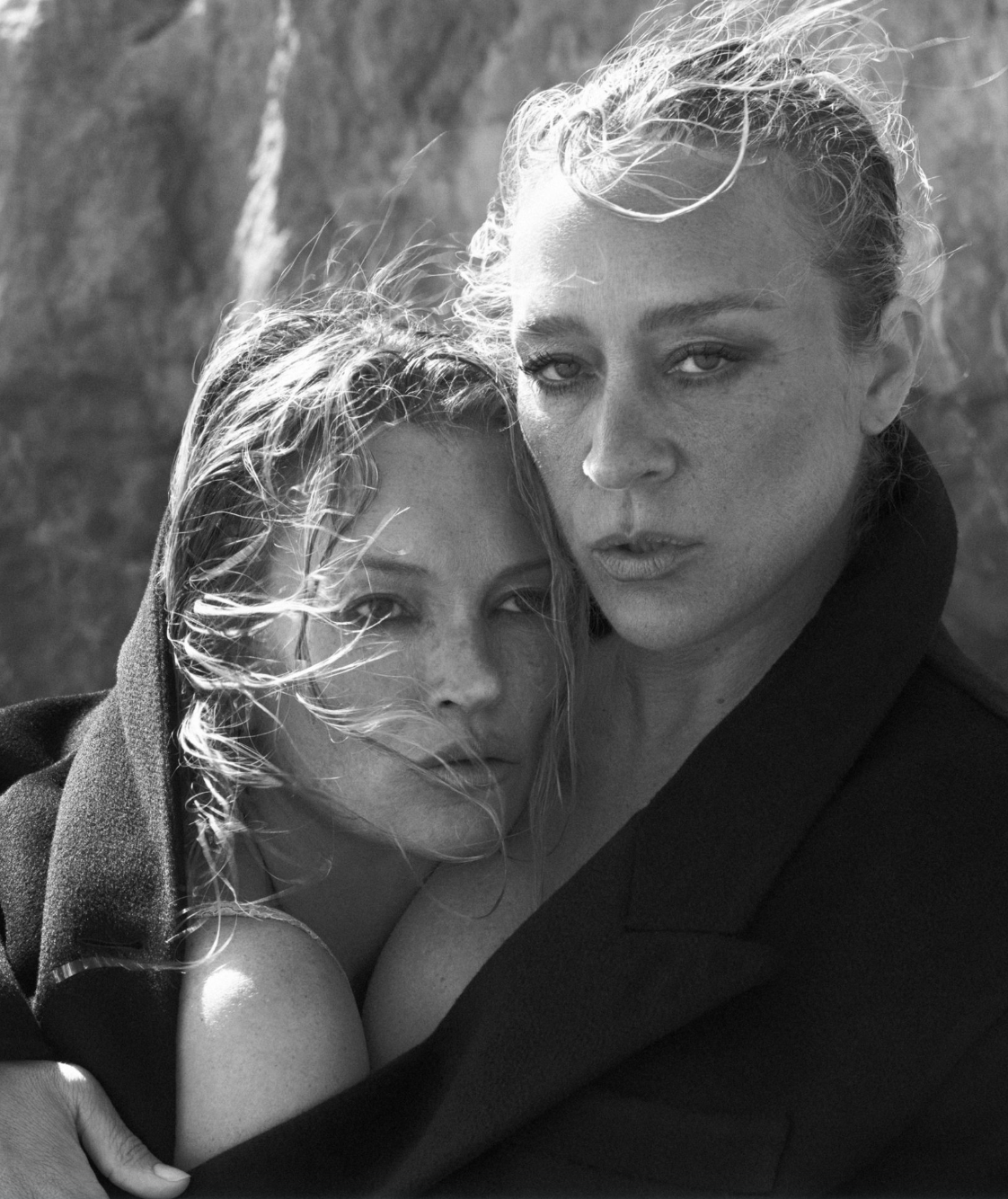 Black and white photograph of two women outdoors, close together, with wind-blown hair. One woman has short, curly hair and is wearing a dark jacket, while the other has long, straight hair partially covered by a dark garment. They are looking direct