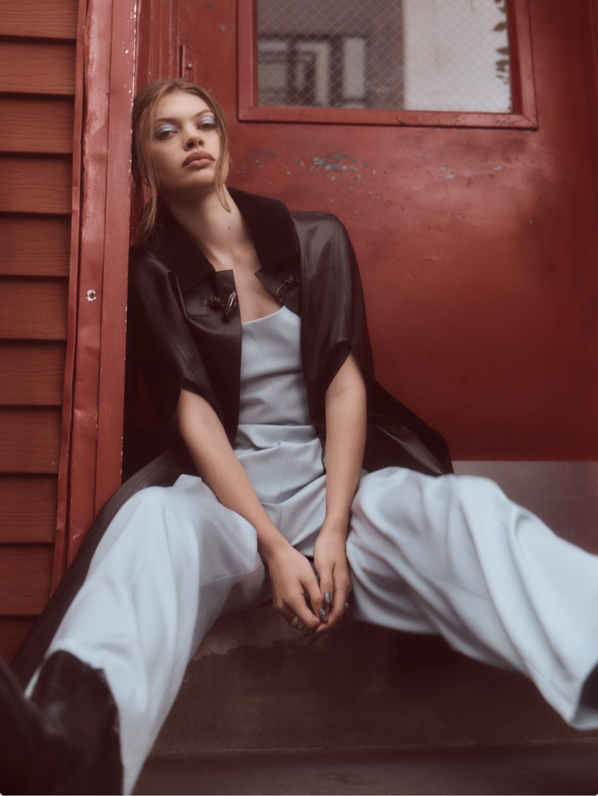 A woman sitting against a red painted wall and door, wearing a black jacket over a white dress with wide-legged pants.