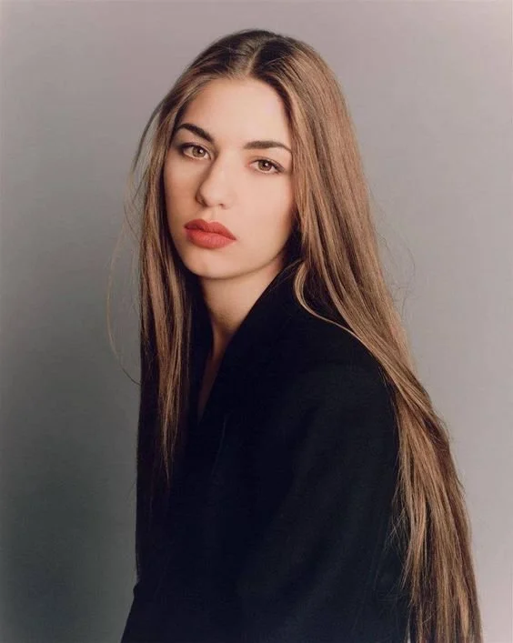 A young woman with long, light brown hair and bold makeup, wearing a black top, looking confidently at the camera against a plain gray background.
