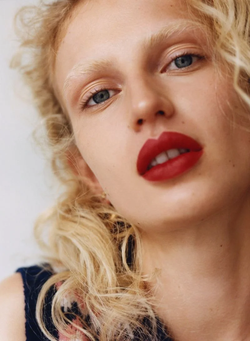 Close-up of a young woman with curly blonde hair, blue eyes, and bold red lipstick, looking at the camera.