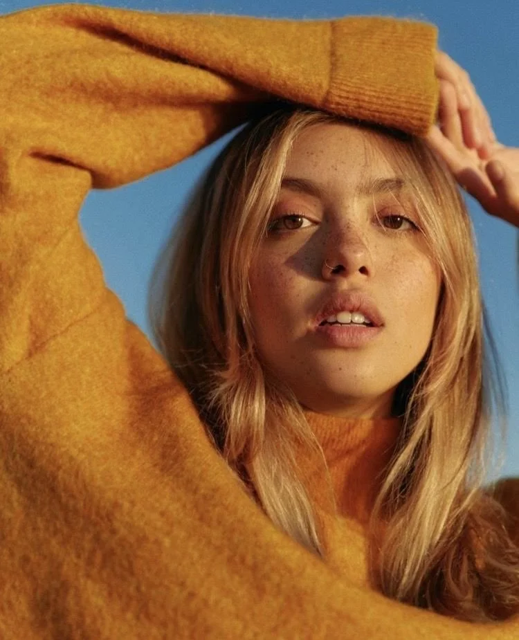 Close-up of a young woman with long blonde hair, freckles, and a nose piercing, wearing a mustard yellow sweater, posing outdoors against a blue sky.