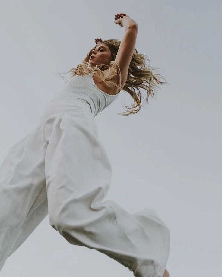 A woman with long, blonde hair wearing a white sleeveless top and wide-legged white pants, looking sideways with her arms raised above her head, against a plain, light gray background.