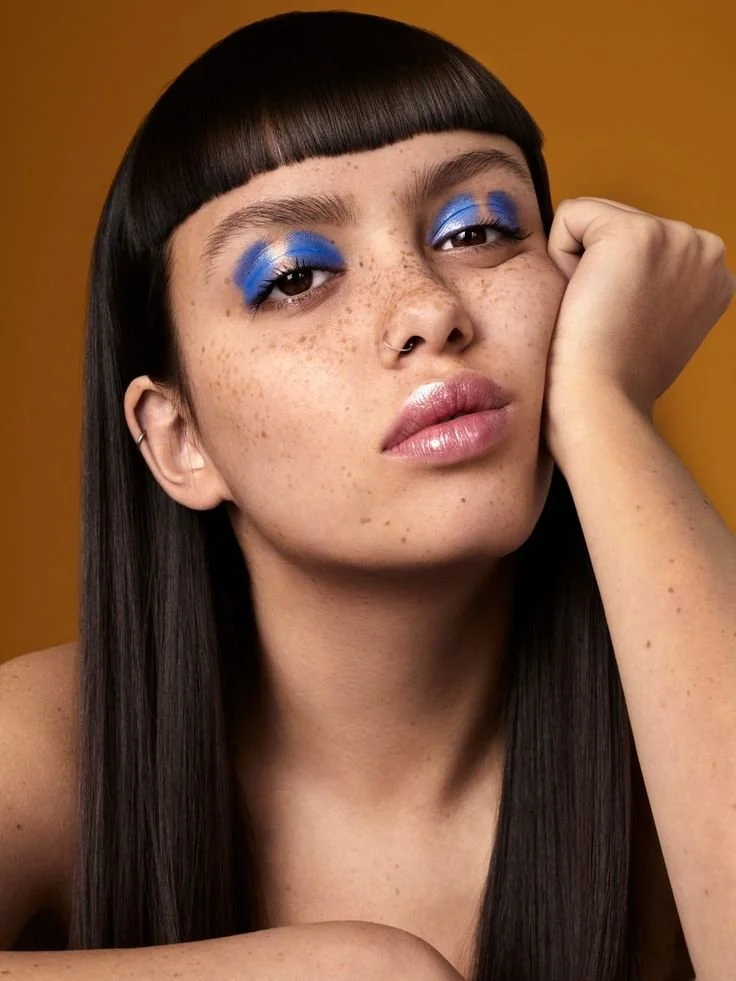 Close-up of a young woman with dark straight hair and freckles, resting her head on her hand, wearing vibrant blue eyeshadow and glossy lips, with a warm brown background.
