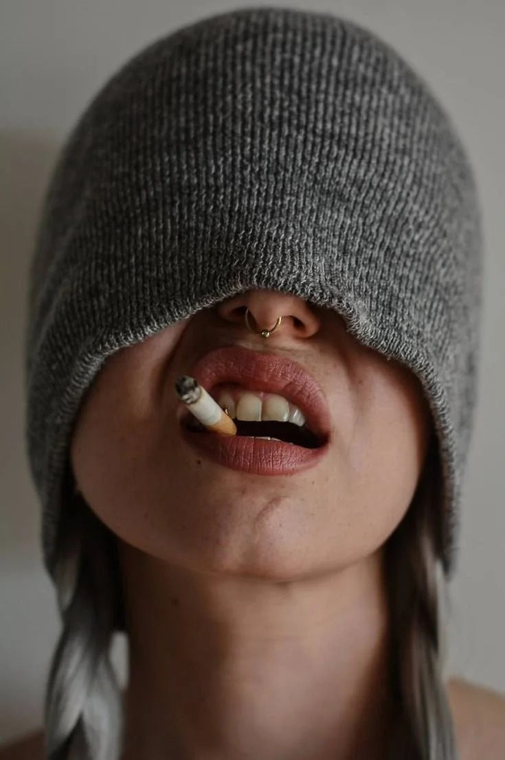 Person with a gray knit hat covering eyes, holding a cigarette in mouth, showing teeth, with a septum piercing.