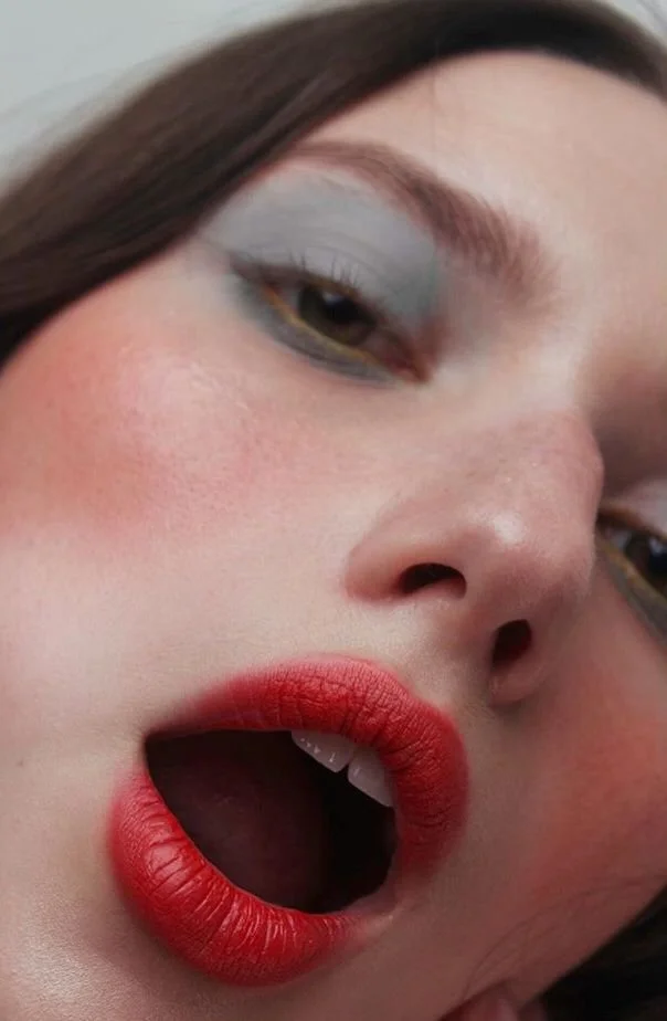 Close-up of a woman's face with bold makeup, including gray eyeshadow, red lipstick, and blush on her cheeks, with her mouth slightly open.