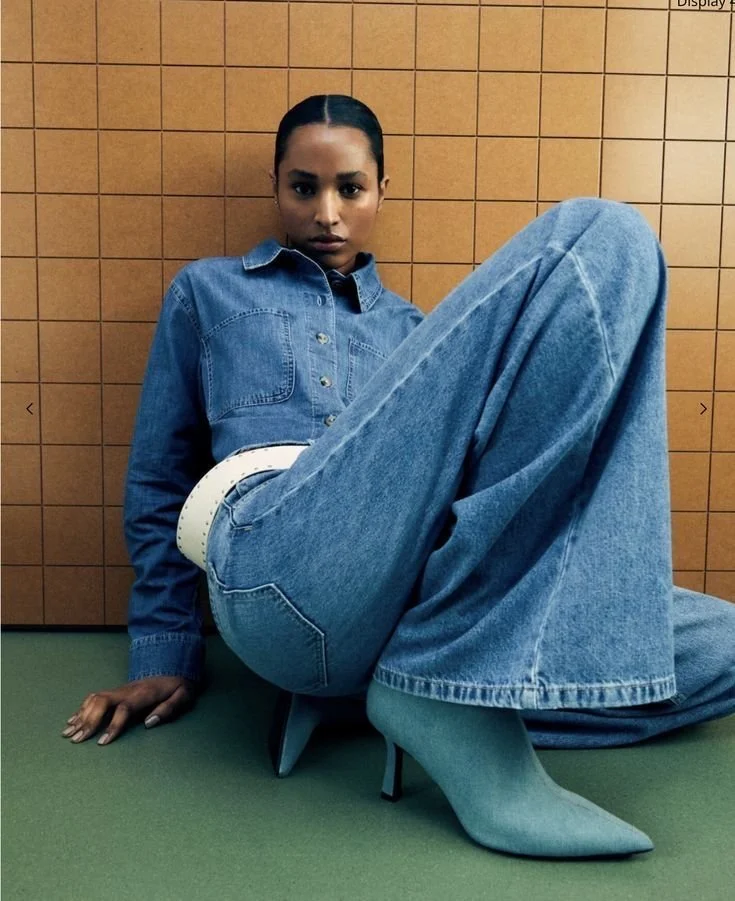 A woman wearing a denim shirt, large denim jeans with a belt, and pointed high heels, sitting against a tan tiled wall with green flooring.