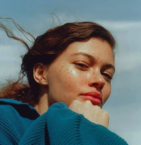 Close-up of a woman with red hair, freckles, and relaxed expression, wearing a blue hoodie and outdoors.