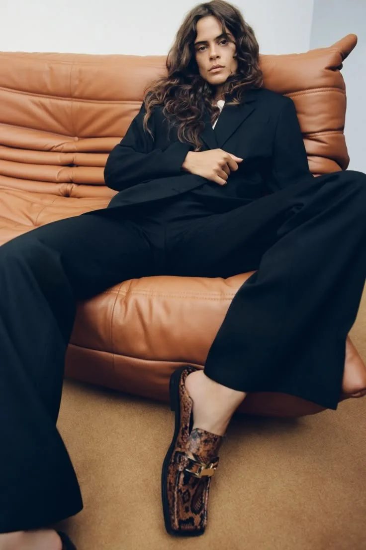 Woman with long wavy hair sitting on a tan leather sofa, wearing a black suit and leopard print shoes with a gold buckle, in a relaxed pose with one leg extended.
