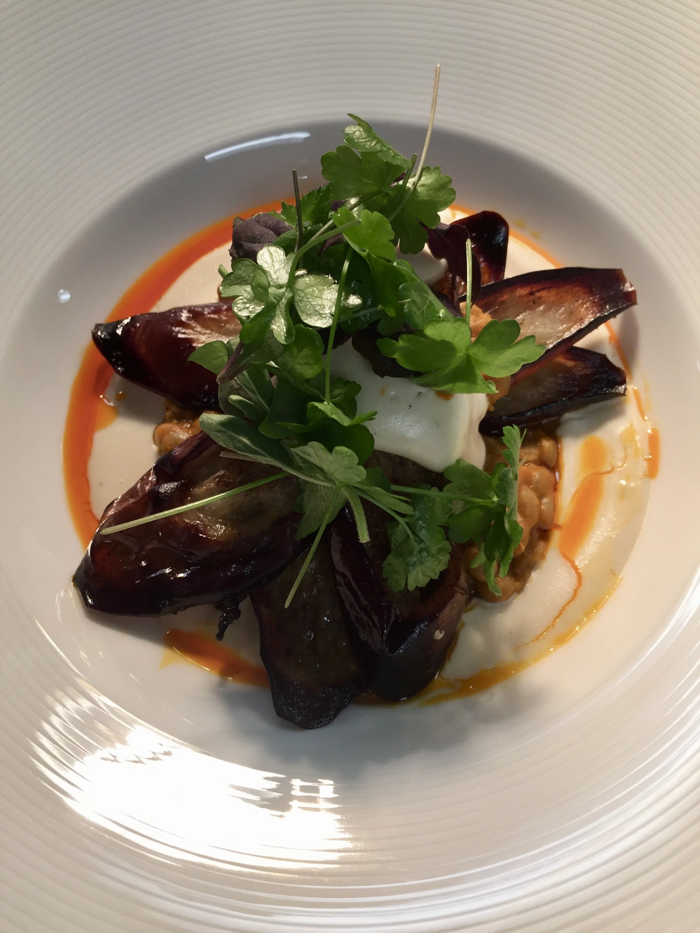 A plated gourmet dish with roasted eggplant, pine-nut cream, fresh herbs garnish, smoked lentil and garbanzos , chilli oil